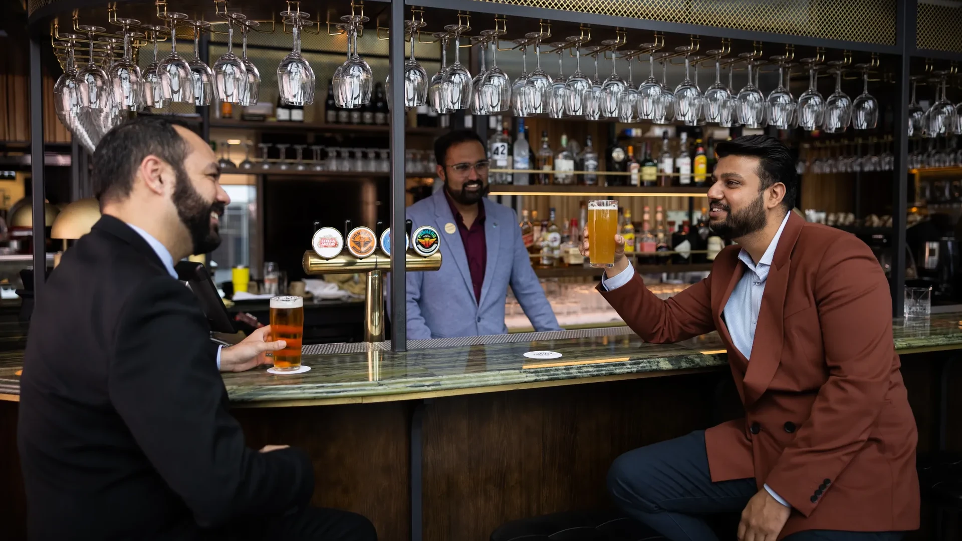 Two men in suits enjoying beers at a bar, chatting with a bartender in a modern, well-stocked setting.