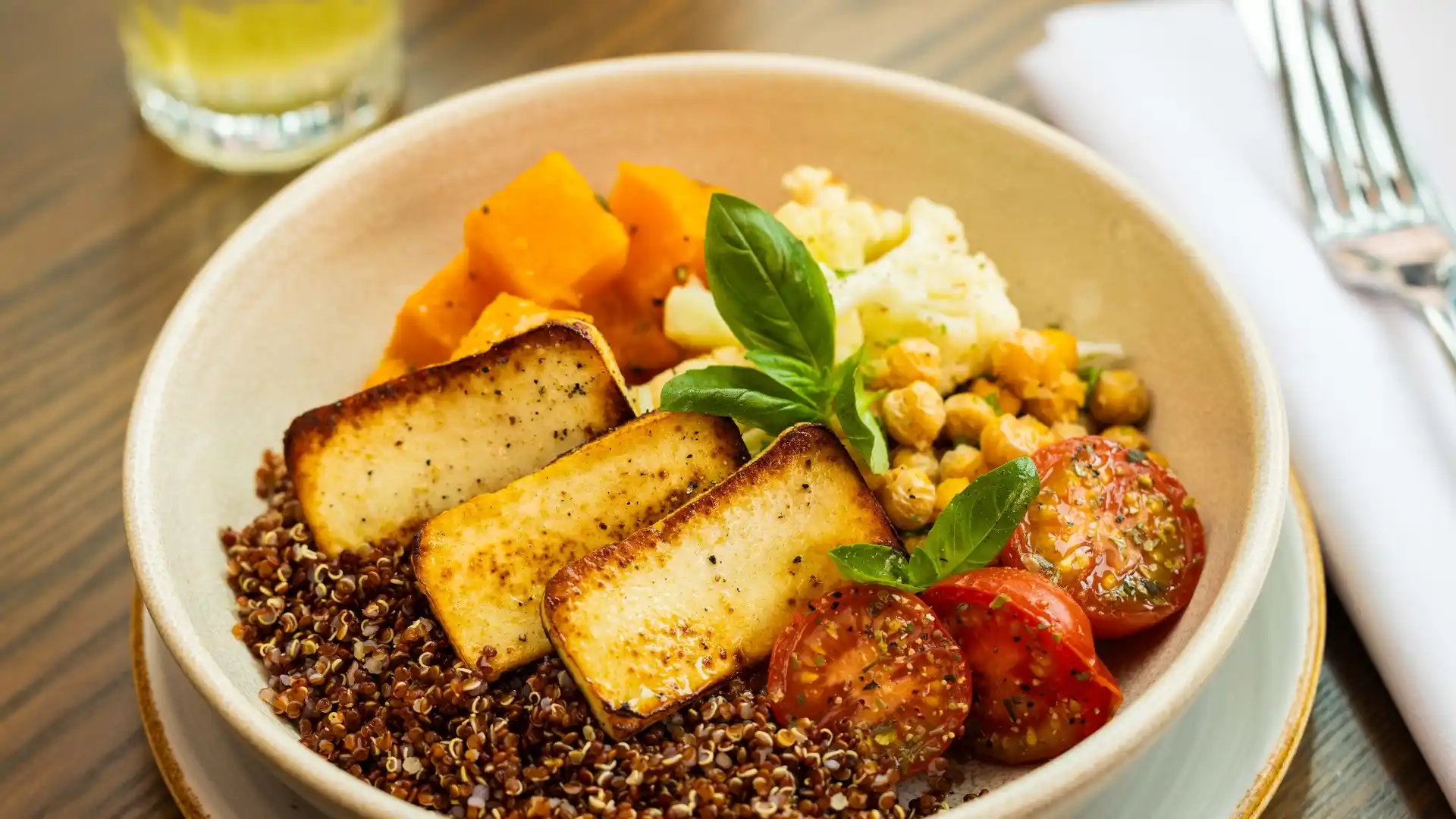 Grain bowl with quinoa, grilled halloumi, roasted pumpkin, chickpeas, cherry tomatoes, and fresh basil, served on a wooden table.