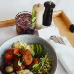 Bowl with falafel, hummus, olives, cherry tomatoes, cucumber, and grains, served with a berry drink and condiments on a tray.