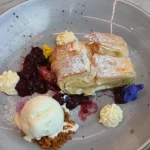 A plated pastry dessert at Thirteen 50 Kitchen & Bar Auckland, dusted with icing sugar and served with berry compote, cream dollops, edible flowers, and a scoop of vanilla ice cream.
