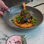 A person holding a bowl at Thirteen 50 Kitchen & Bar Auckland featuring a braised lamb shank on creamy orange mash, topped with green peas and herbs.