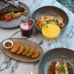 A marble table at Thirteen 50 Kitchen & Bar Auckland featuring a steak being cut with a fork and knife, alongside a bowl of pasta, crispy croquettes with dipping sauce, a pan-seared fish dish, and two cocktails—one bright yellow with lime and one deep red garnished with a blood orange slice.