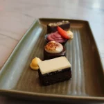 Selection of bite-sized desserts including layered chocolate cake, cream-topped pastries, and fruit garnishes arranged on a rectangular plate.