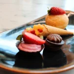 Assorted desserts including a mini doughnut topped with a strawberry, a filled cone with diced fruit, a strawberry macaron, and a chocolate praline on a plate.