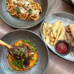 Overhead view of a table with a lamb shank on mash with peas, a pasta dish topped with parmesan and herbs, and a pulled pork sandwich with fries and ketchup.