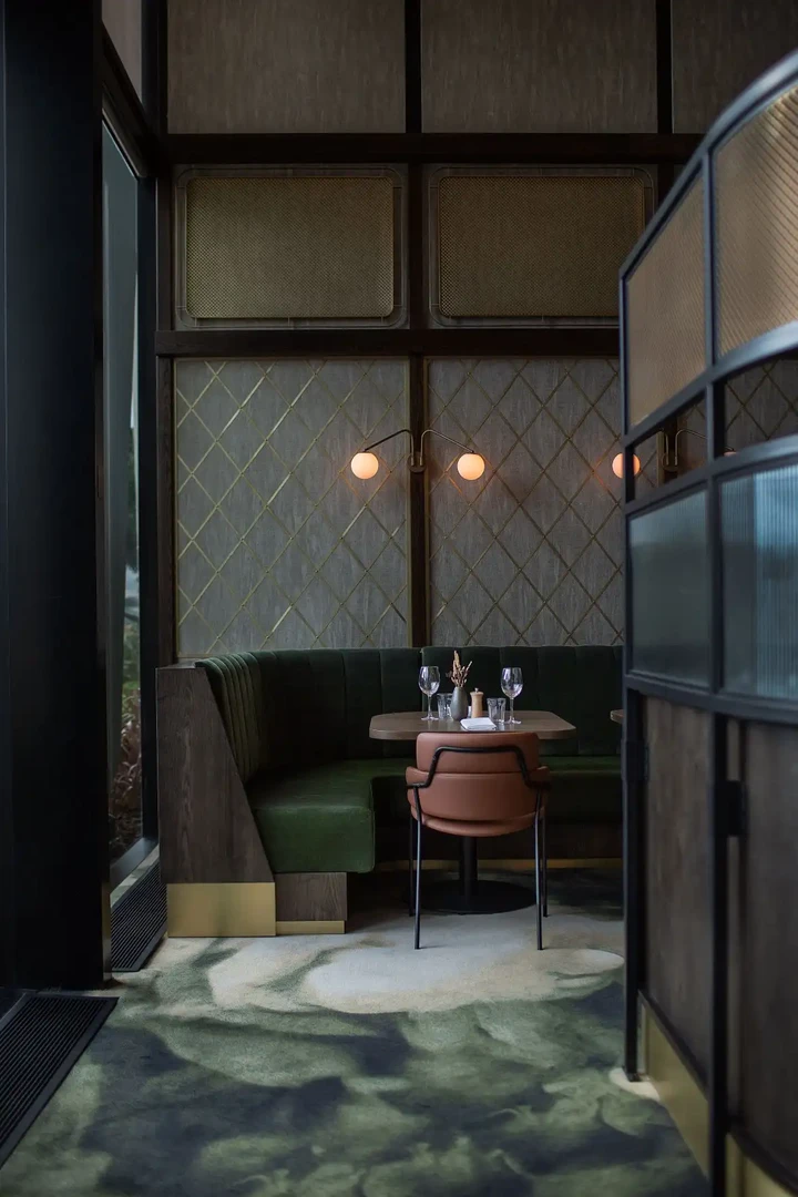 A cozy, modern restaurant booth set for two. There’s a curved green upholstered bench, a small round table with wine glasses and tableware, and a single chair opposite. The space has warm ambient lighting from wall-mounted globe lights, dark wood paneling, and a stylish partition on the right, giving it a semi-private, intimate dining feel.