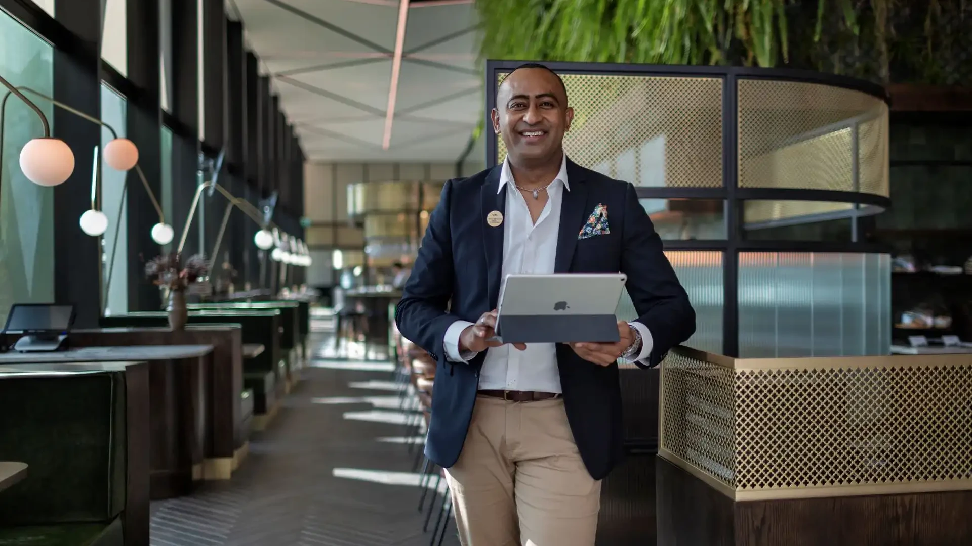 Smiling restaurant manager in a suit holding a tablet, standing inside a modern dining space with booths and pendant lighting.