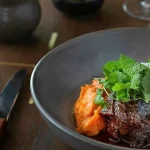 Braised beef in rich sauce served with mashed sweet potato and fresh herbs, plated in a bowl on a wooden table.